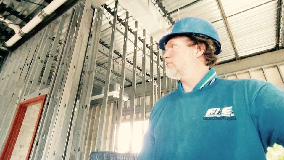 An F.I.E. Electric electrician in a blue hard hat surveying a commercial build-out framed in steel studs